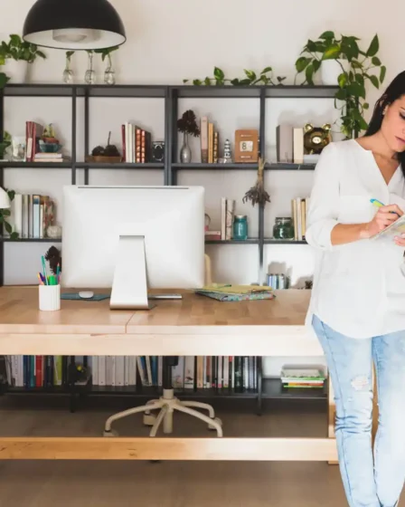 Imagem de um espaço de home office com uma mulher escrevendo em um bloco de notas, mesa organizada, livros na prateleira e plantas decorativas, ideal para montar um cantinho funcional de baixo custo para leitura ou trabalho.