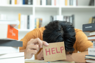 Pessoa com a cabeça apoiada sobre a mesa segura cartão com a palavra “HELP”, cercada por pilhas de livros em ambiente de estudo.