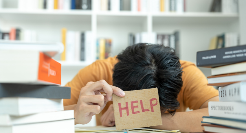 Pessoa com a cabeça apoiada sobre a mesa segura cartão com a palavra “HELP”, cercada por pilhas de livros em ambiente de estudo.