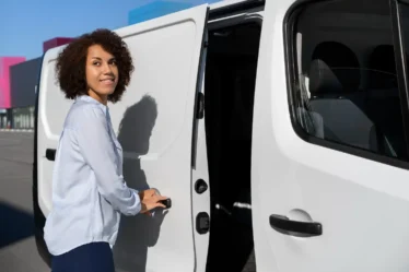 Mulher sorridente preparando van branca para transporte de convidados em evento, destacando a locação de van para transporte de convidados com conforto e logística inteligente.