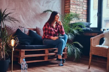 Estilo Industrial na decoração: Como aplicar sem deixar o ambiente frio: Mulher sentada em um sofá de madeira em um ambiente aconchegante com parede de tijolos à vista, plantas e luminária decorativa, estilo industrial na decoração com elementos quentes que evitam o ambiente frio.