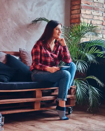 Estilo Industrial na decoração: Como aplicar sem deixar o ambiente frio: Mulher sentada em um sofá de madeira em um ambiente aconchegante com parede de tijolos à vista, plantas e luminária decorativa, estilo industrial na decoração com elementos quentes que evitam o ambiente frio.