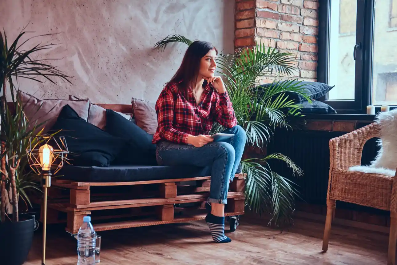 Estilo Industrial na decoração: Como aplicar sem deixar o ambiente frio: Mulher sentada em um sofá de madeira em um ambiente aconchegante com parede de tijolos à vista, plantas e luminária decorativa, estilo industrial na decoração com elementos quentes que evitam o ambiente frio.