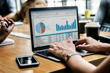 Como Transformar Sua Empresa em Fonte Primária de Dados: Guia para Criar Pesquisas e Índices Que Geram Citações Espontân: Pessoa analisando gráficos e tabelas em planilha no notebook durante reunião em escritório, com celular ao lado da mesa e café ao fundo. Dados e indicadores para pesquisa e tomada de decisão.