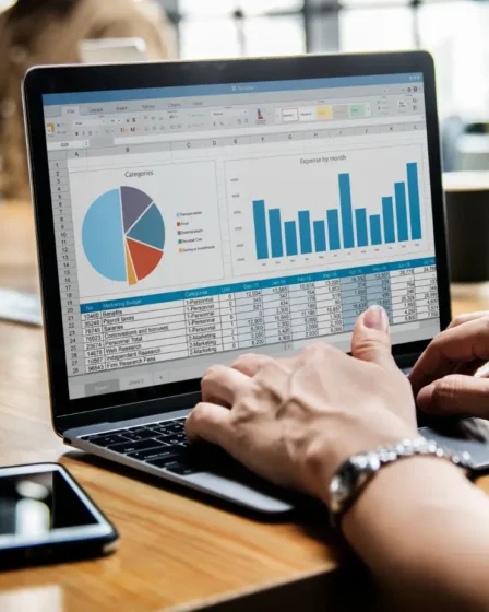 Como Transformar Sua Empresa em Fonte Primária de Dados: Guia para Criar Pesquisas e Índices Que Geram Citações Espontân: Pessoa analisando gráficos e tabelas em planilha no notebook durante reunião em escritório, com celular ao lado da mesa e café ao fundo. Dados e indicadores para pesquisa e tomada de decisão.