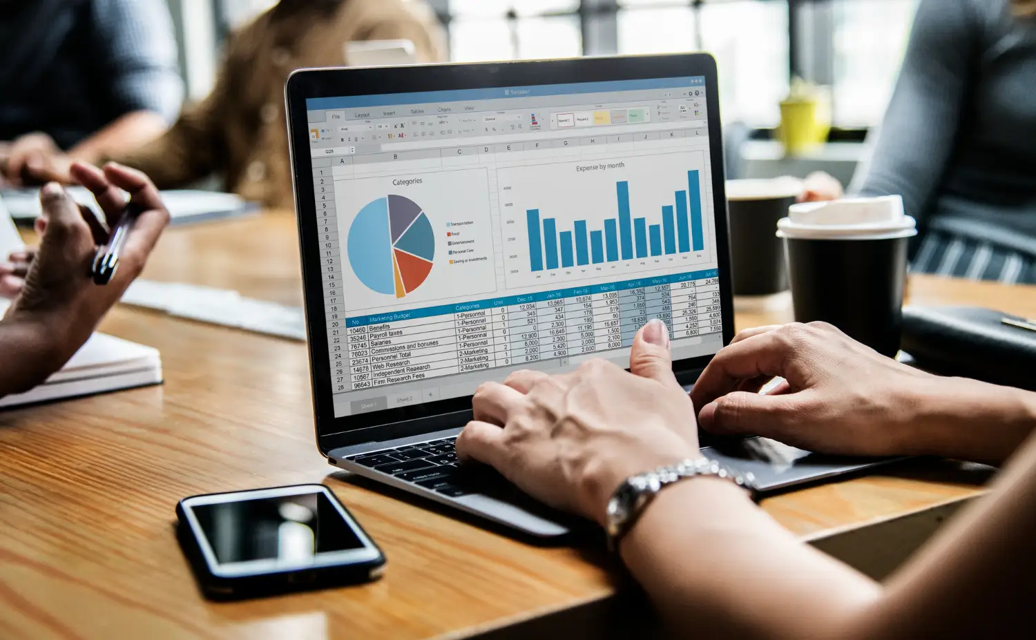 Como Transformar Sua Empresa em Fonte Primária de Dados: Guia para Criar Pesquisas e Índices Que Geram Citações Espontân: Pessoa analisando gráficos e tabelas em planilha no notebook durante reunião em escritório, com celular ao lado da mesa e café ao fundo. Dados e indicadores para pesquisa e tomada de decisão.